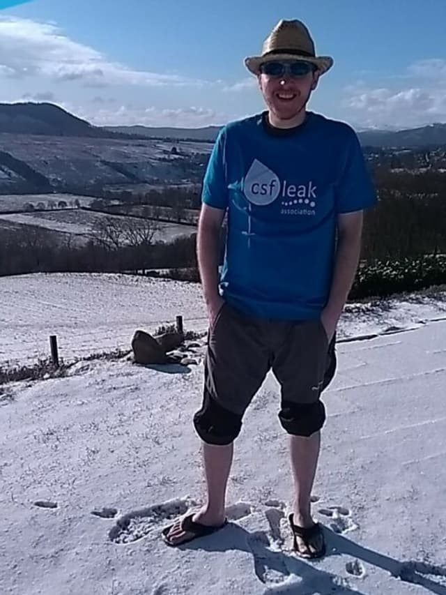 A smiling man wearing a hat, sunglasses, and a CSF Leak Association shirt, and flip flops in the snow outside.