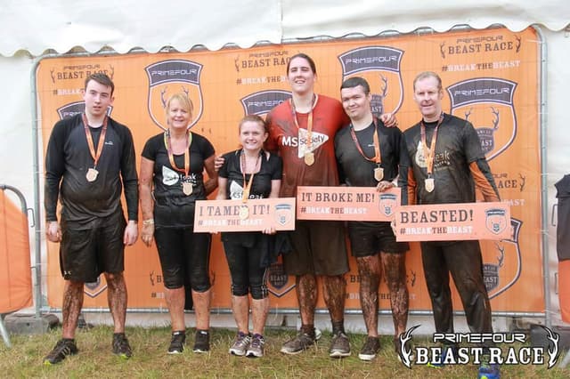 A group of people with muddy legs and wearing medals in front of the backdrop for the Beast Race.