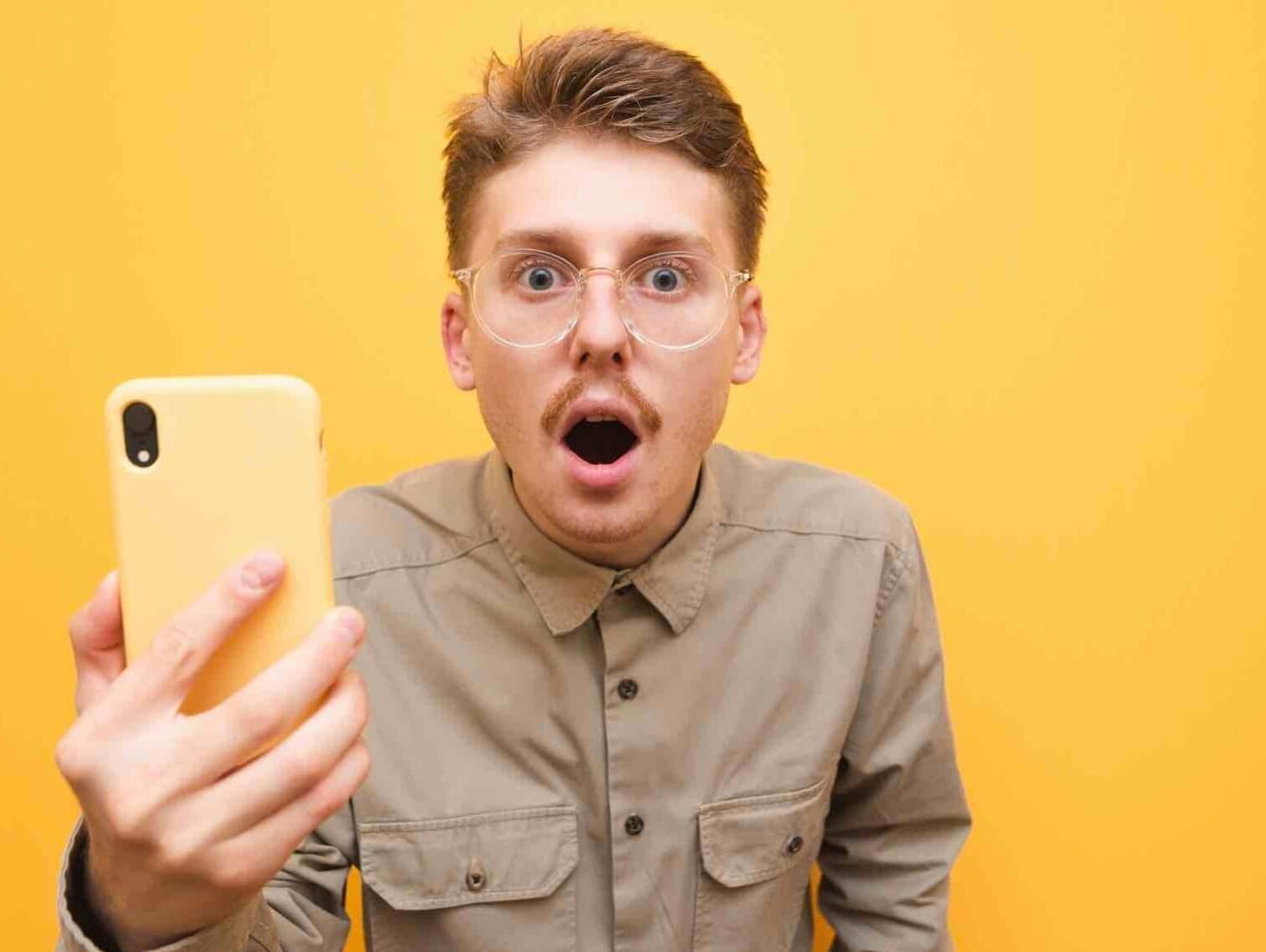 Mann mit roten Haaren, Brille und Schnurrbart hält gelbes Smartphone ist entsetzt