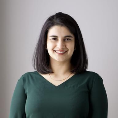 Photograph of Faryal shehzad, she is wearing a green top and a gold necklace, and smiling against a plain grey backdrop.