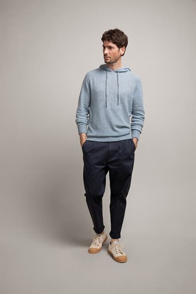Johnstons of Elgin's Denim Blue Gauzy Knit Cashmere Hoodie on model wearing navy trousers with cream trainers on a grey background