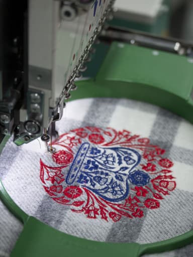 Johnstons of Elgin Grey Check Double Face Lambswool Throw, being embroidered in the red and blue of the coronation emblem.