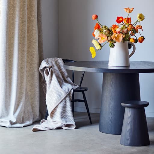 Johnstons of Elgin's Herringbone Jacquard Blanket Stitch Throw in Natural on a chair next to round grey table with a vase of orange and yellow flowers