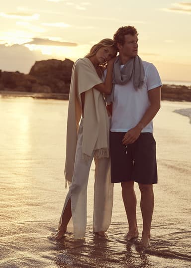 A couple wearing Johnstons of Elgin Summer accessories, female is wearing Natural Cashmere Stole draped over a her shoulder, he is wearing a lightweight silver grey merino scarf wrapped around his neck on a beach at sunset