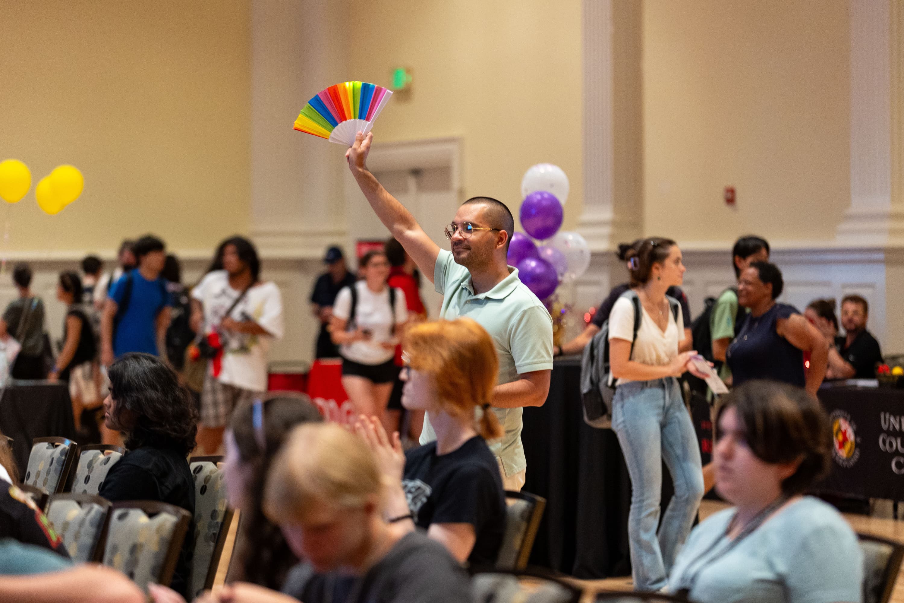 A Quelcome attendee holding up a colorful fan and smiling while other attendees browse the resource tables in the background.