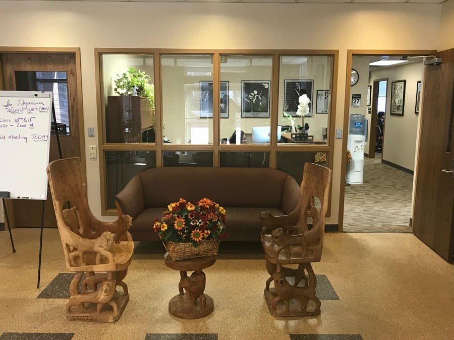 Lobby area with wide cushioned couch, a small carved wooden table with a flower arrangement flanked by intricately carved wood chairs.
