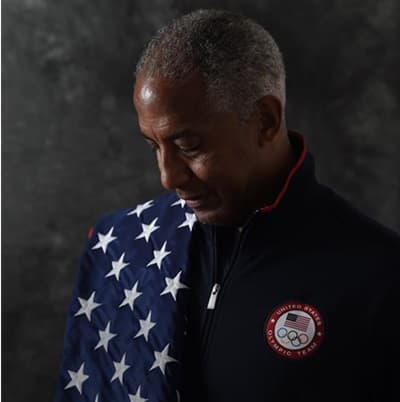 Renaldo Nehemiah wearing a US olympic team patch with a flag over one shoulder, looking down