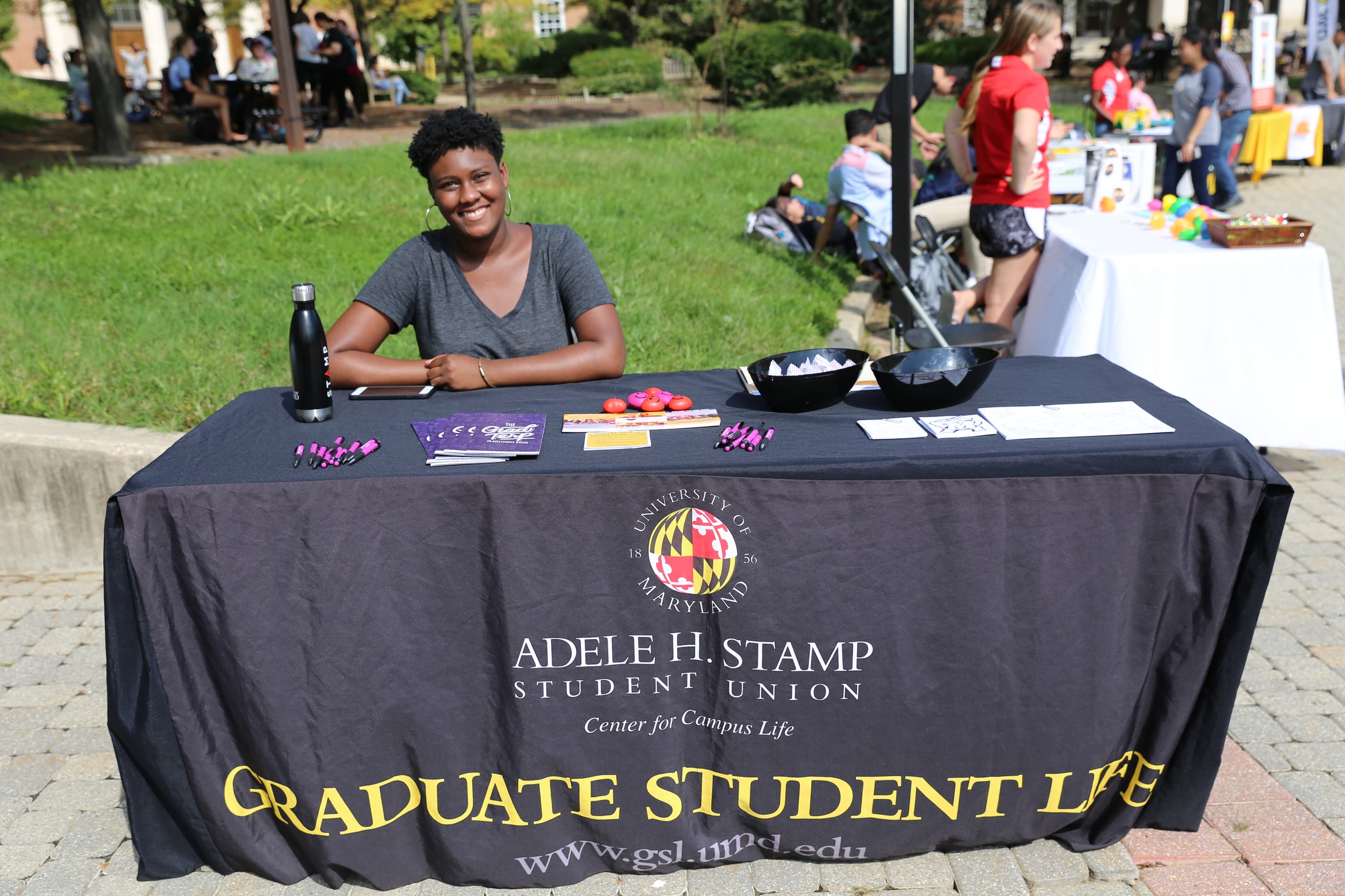 Black woman smiling at the camera, sitting beside a table with papers and pens on top. Tablecloth says Adele H. Stamp Student Union Center for Campus Life: Graduate Student Life