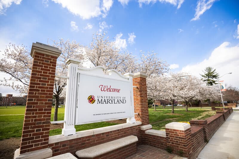 Sign at the entrance of the university that says Welcome, followed by the university logo. The white sign is sandwiched between two brick pillars.