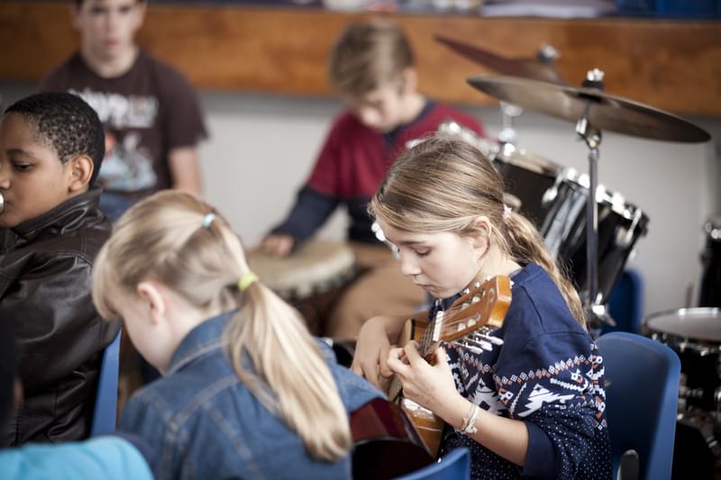 A group of children playing djembe, drum kit, and acoustic guitars.