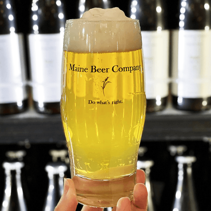 A pint of beer held up in front of a cooler filled with bottles of beer. The glass has the Maine Beer Company logo on it.
