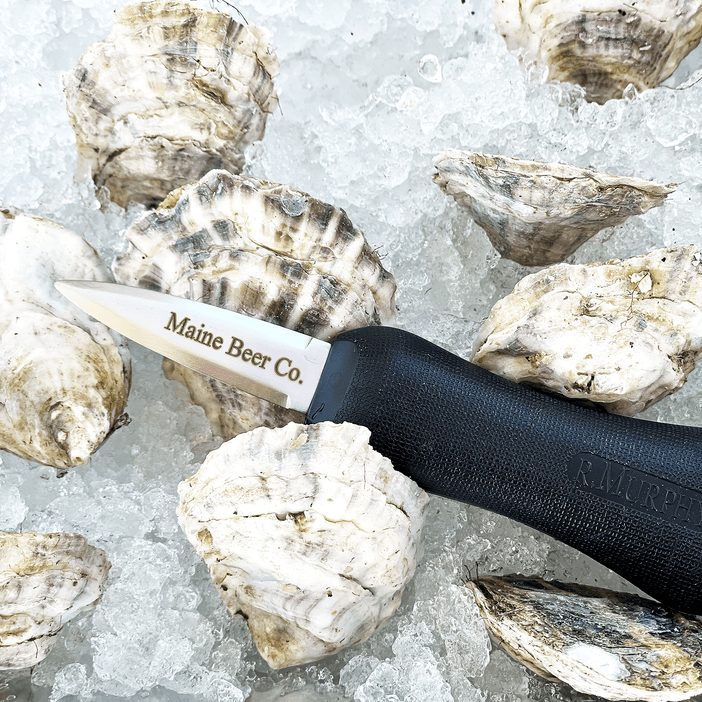 An oyster knife with "Maine Beer Co." engraved on it, sits on a bed of ice and oysters.
