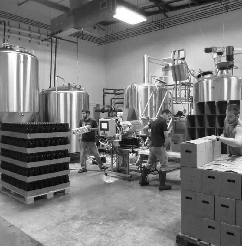 A black and white photo of the early Portland brewery space of MBC with three employees brewing and packaging.
