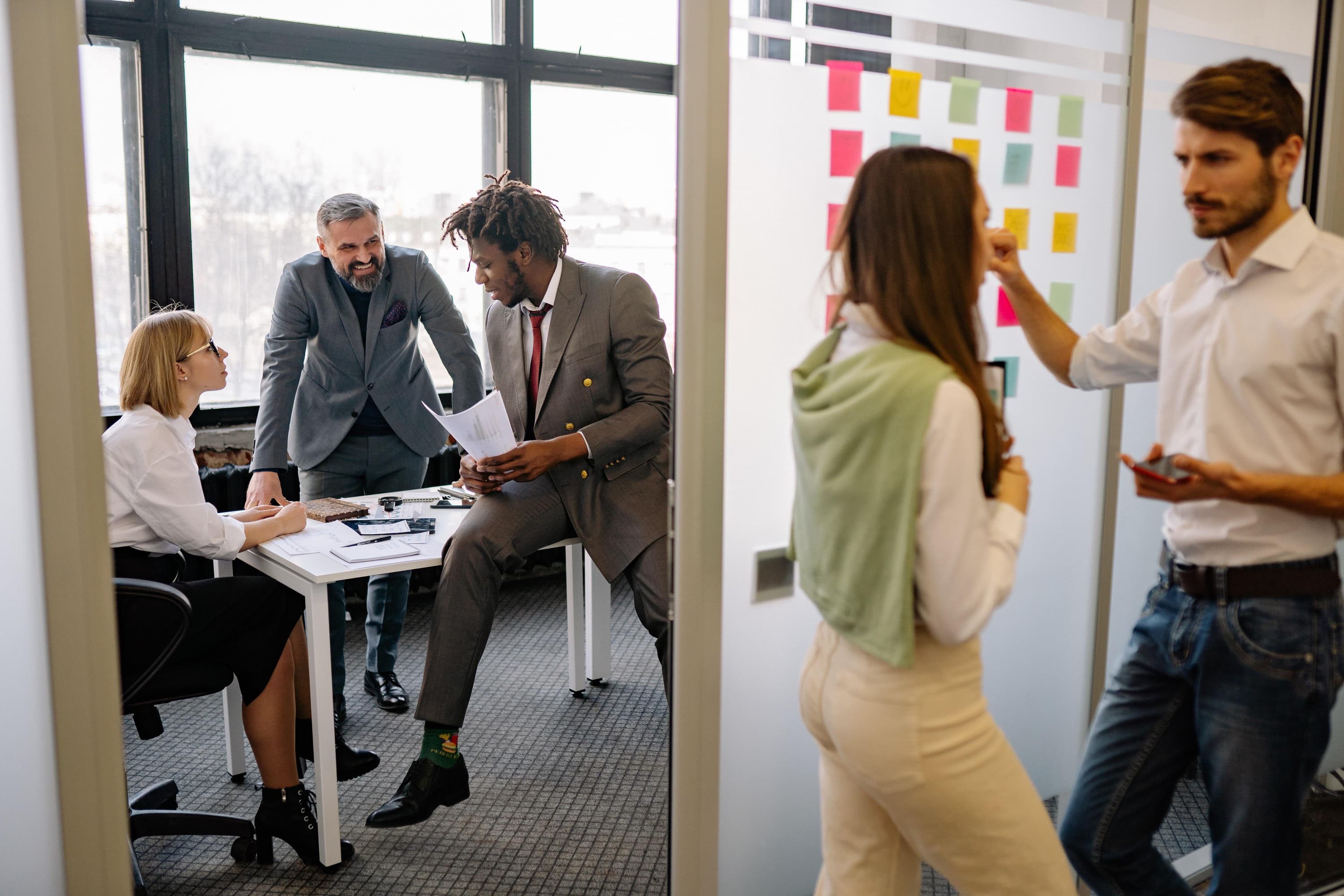 Five working employees, two talking over next to a sticky-note board while the other three talk over some documents on a table.