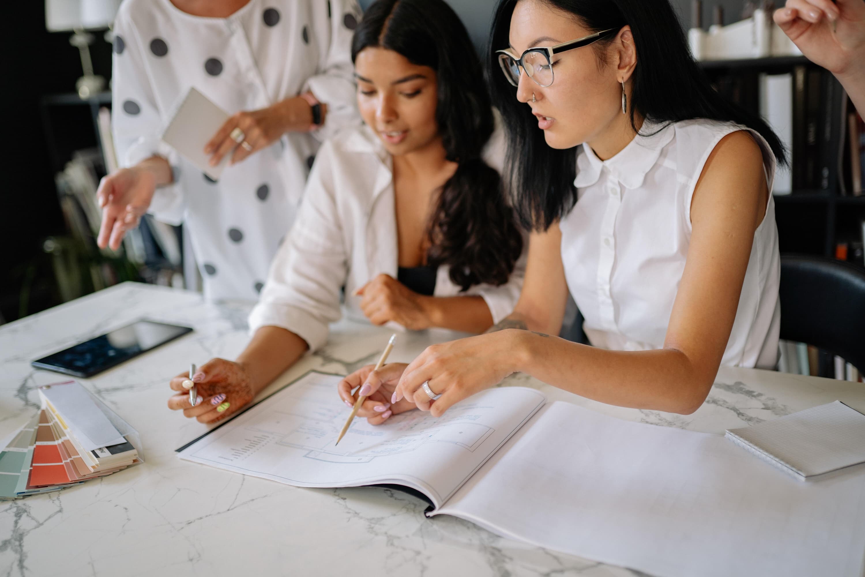Female coworkers talk over designs and paint samples, each wearing a white blouse.