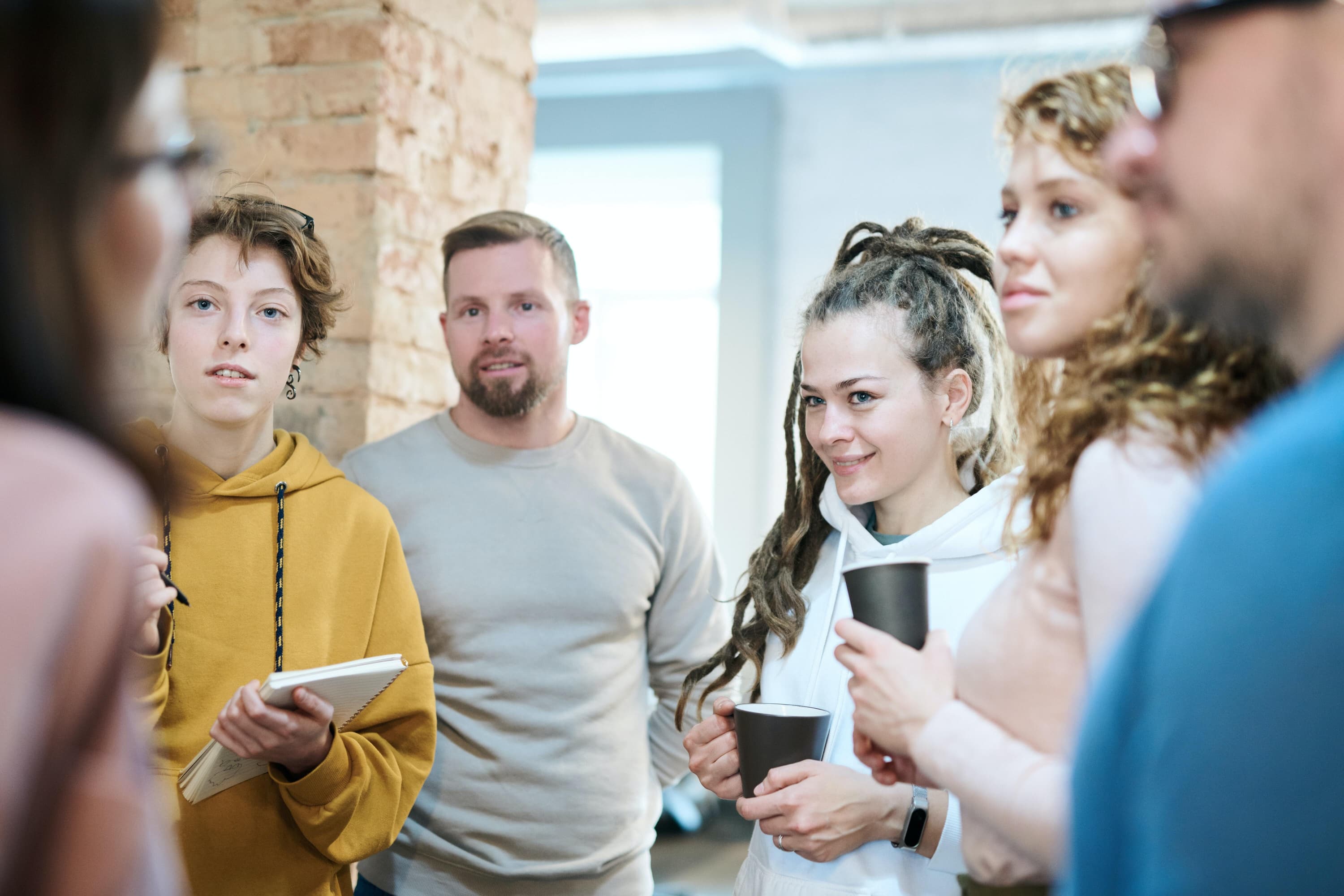 Group of coworkers gathered in a circle talking to each other, two holding coffee cups and one holding a notepad.