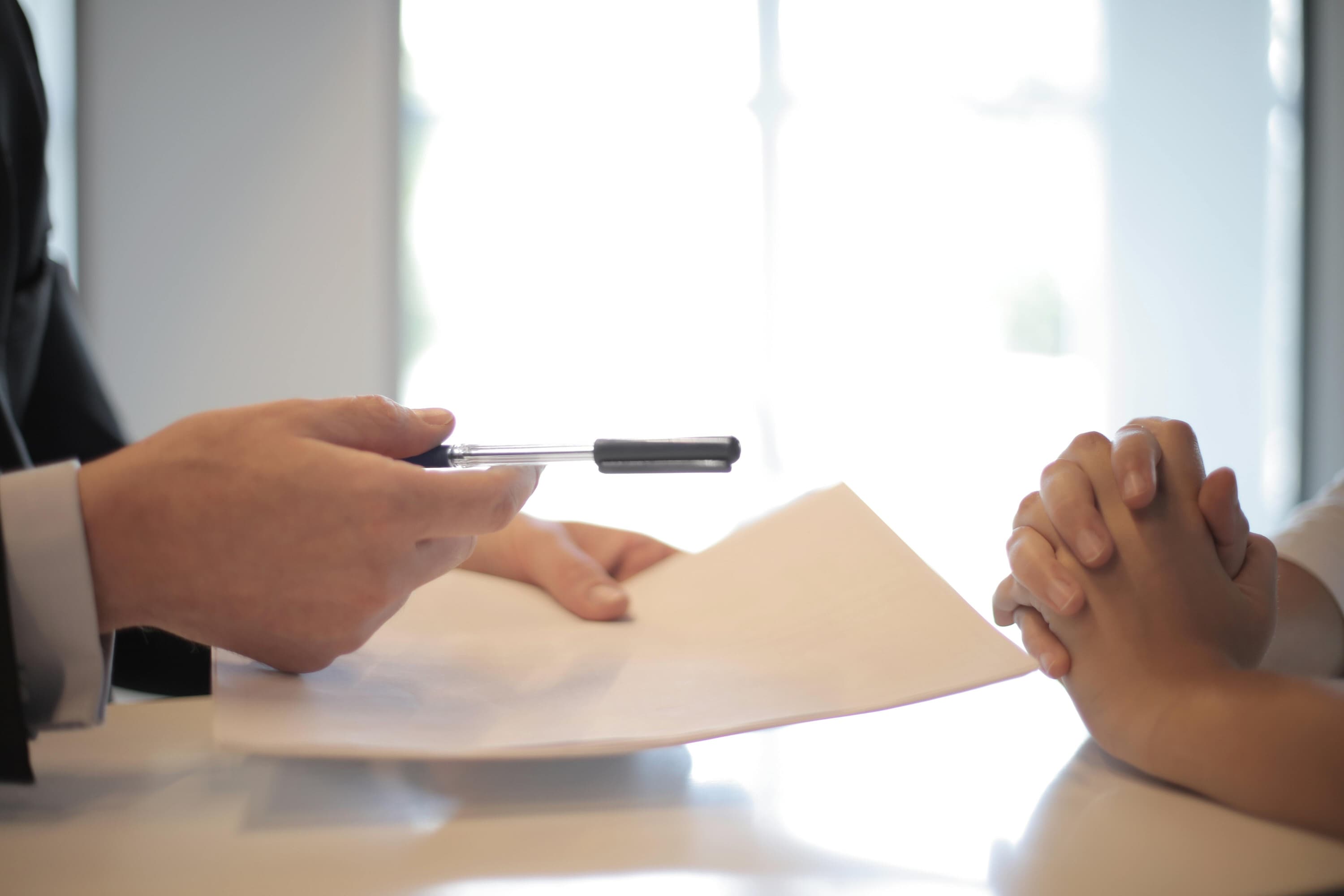 Two people’s hands are pictured, sitting on either side of a table, with one person handing the other forms and a pen.