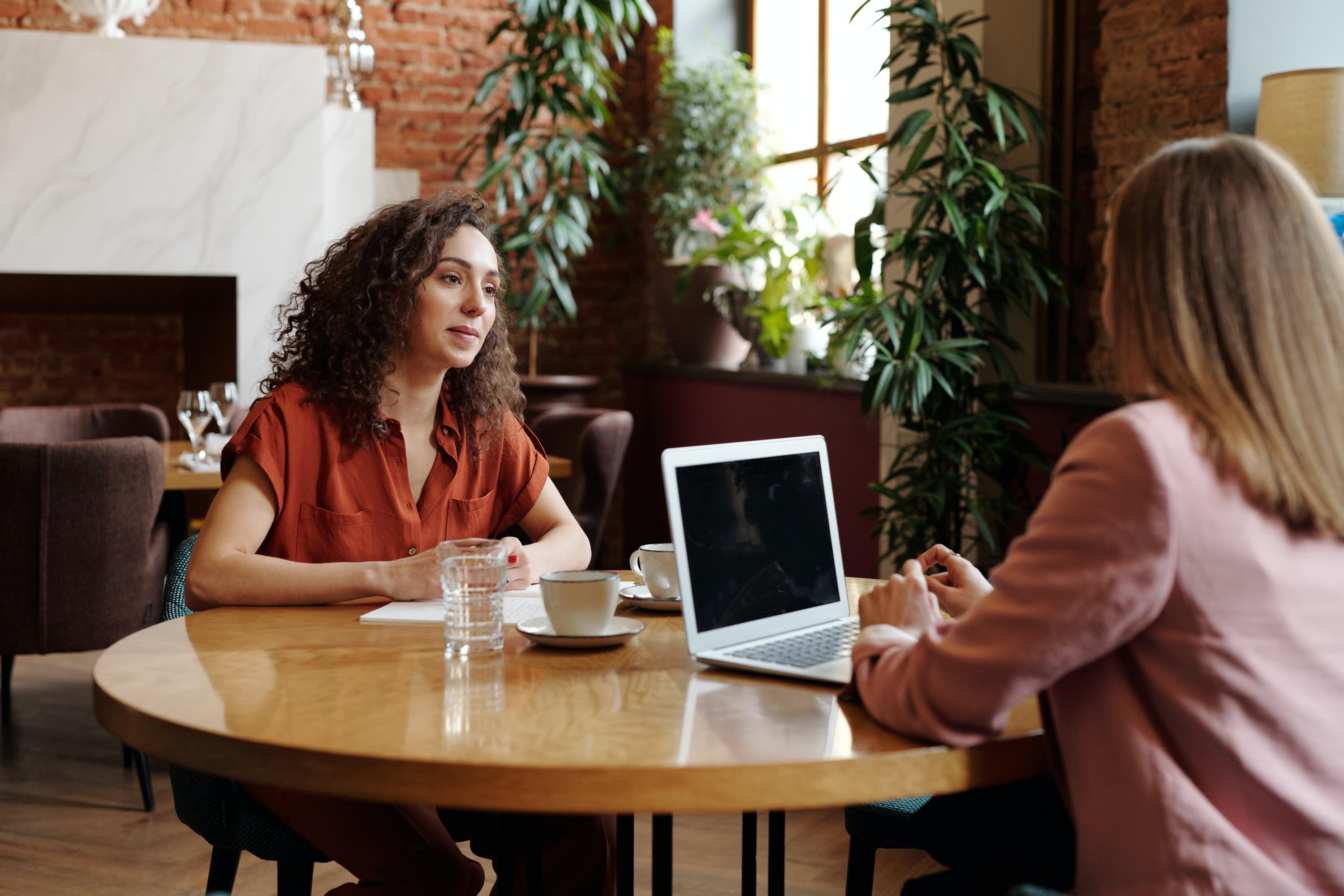 Two people are sitting at a round table drinking coffee and one has their computer open.