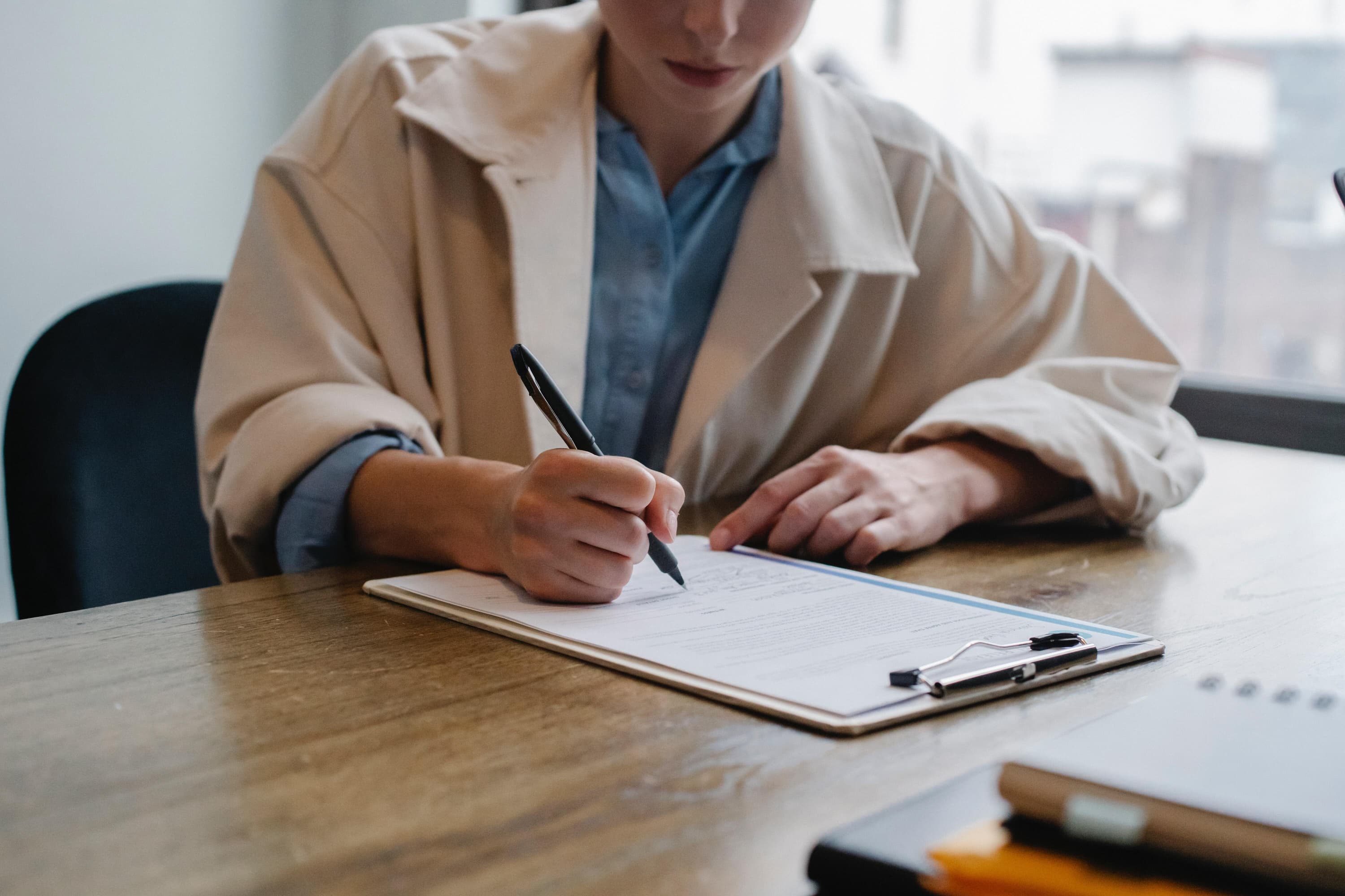 A person wearing a cream jacket and blue button-down is filling out forms on a clipboard with a pen.
