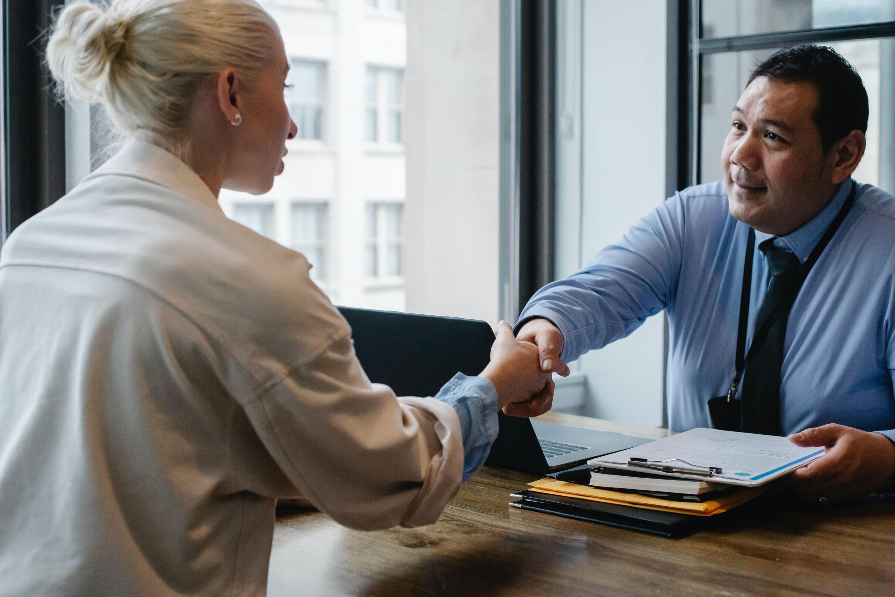 A person wearing a blue button-down and a person wearing a white shirt are shaking hands over a table.