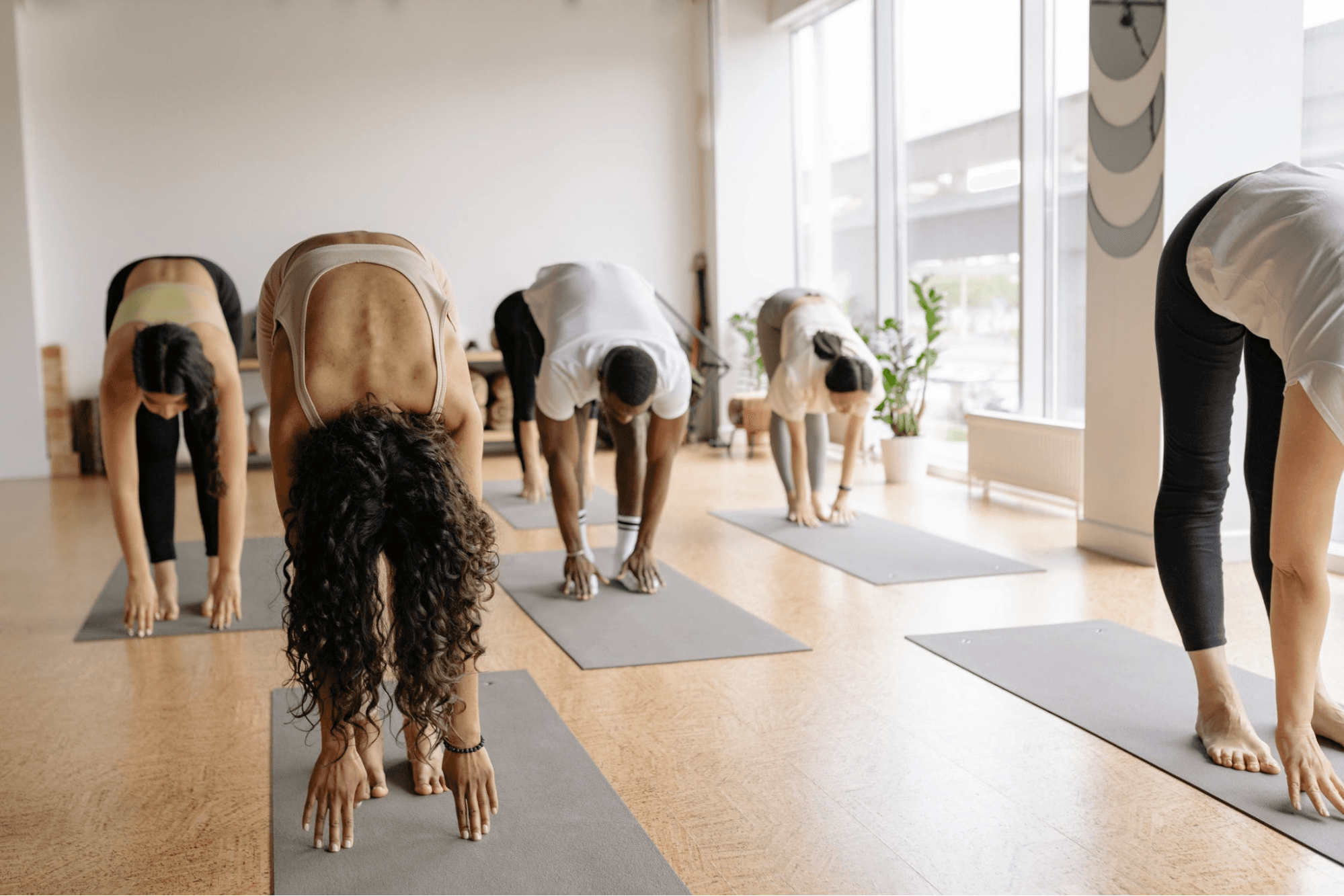 A group of five people are doing yoga in a spacious, light-filled studio. They are all on gray yoga mats and in workout clothes while they are stretching.