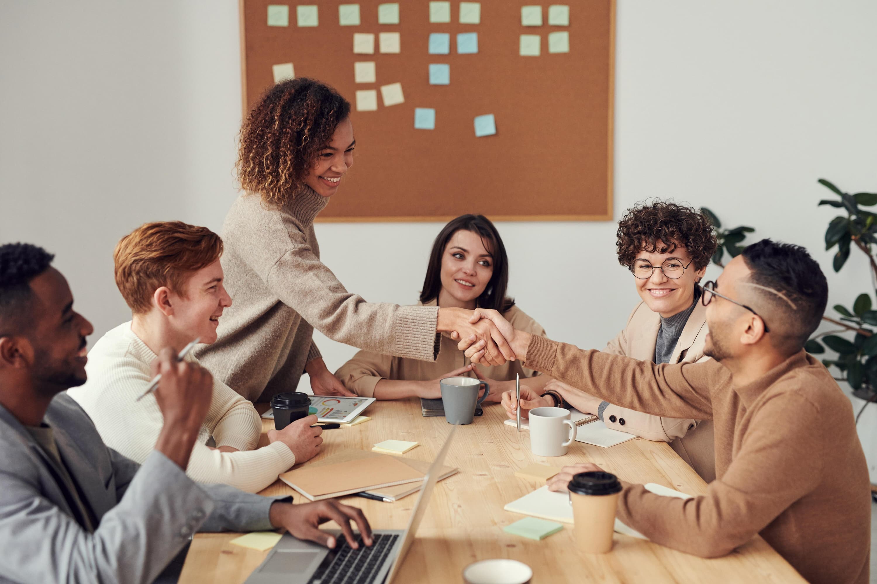 Two co-workers meeting and shaking hands over a table, while other employees are taking notes and drinking coffee.