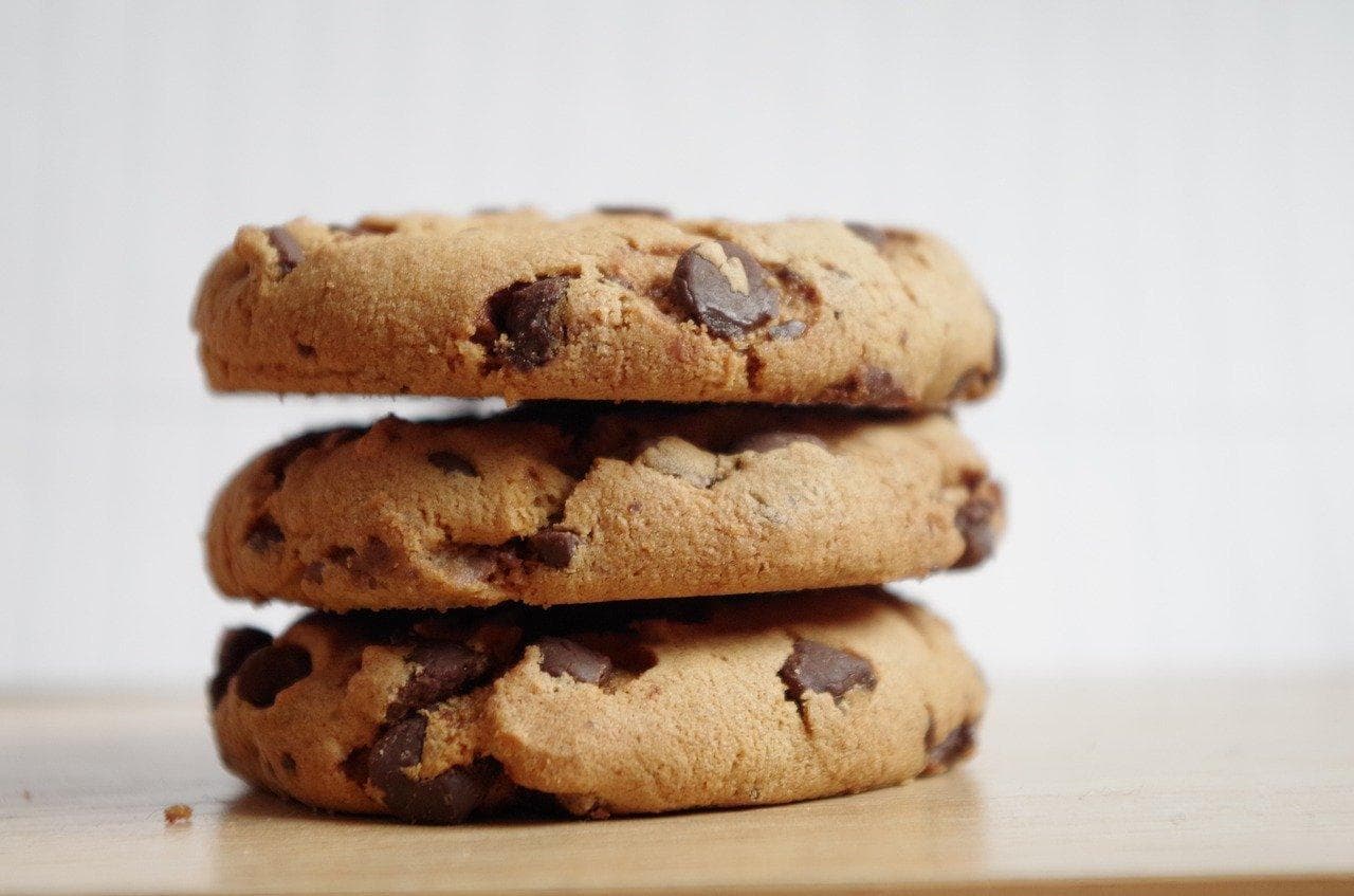 Three chocolate chip cookies are stacked on top of one another in a close-up shot on a tan tabletop.