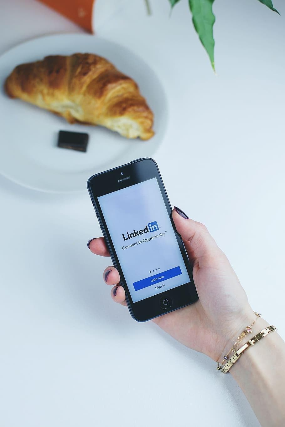 A person is holding a smartphone with the Linkedin app open. They are wearing two gold bracelets, and there is a croissant with chocolate on a plate in front of them.