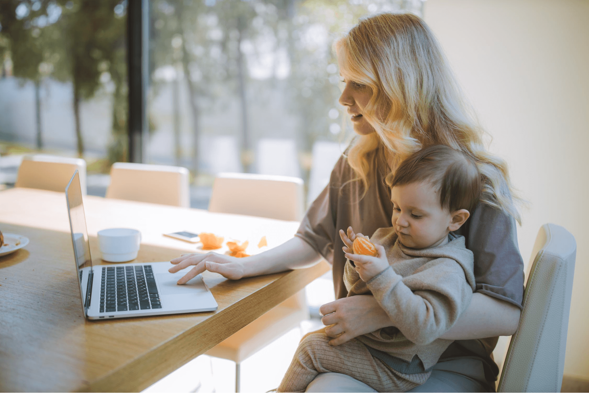 A person is sitting at a tan desk working on their laptop. They are holding their child and drinking a cup of coffee. The child is eating a peeled orange.