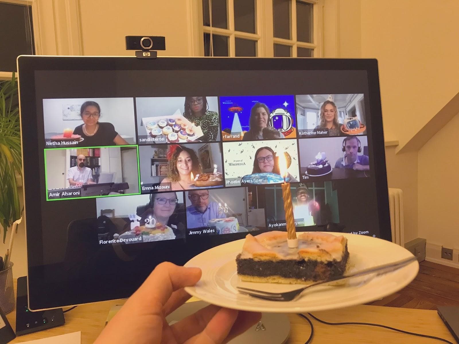 Someone is holding a birthday brownie with a candle on a white plate. They are celebrating a birthday virtually with 11 other people on Zoom.