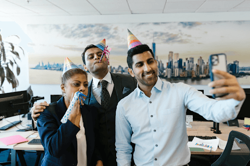 Three people are gathered at an office party and are taking a selfie while wearing party hats.