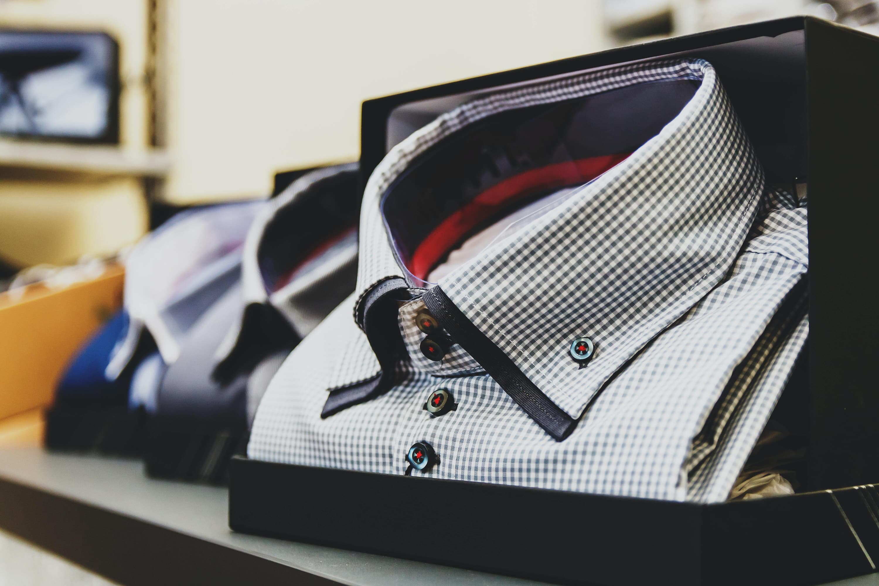 A close-up image of a box with a checkered button-up shirt. There are two other shirts shown in the background.