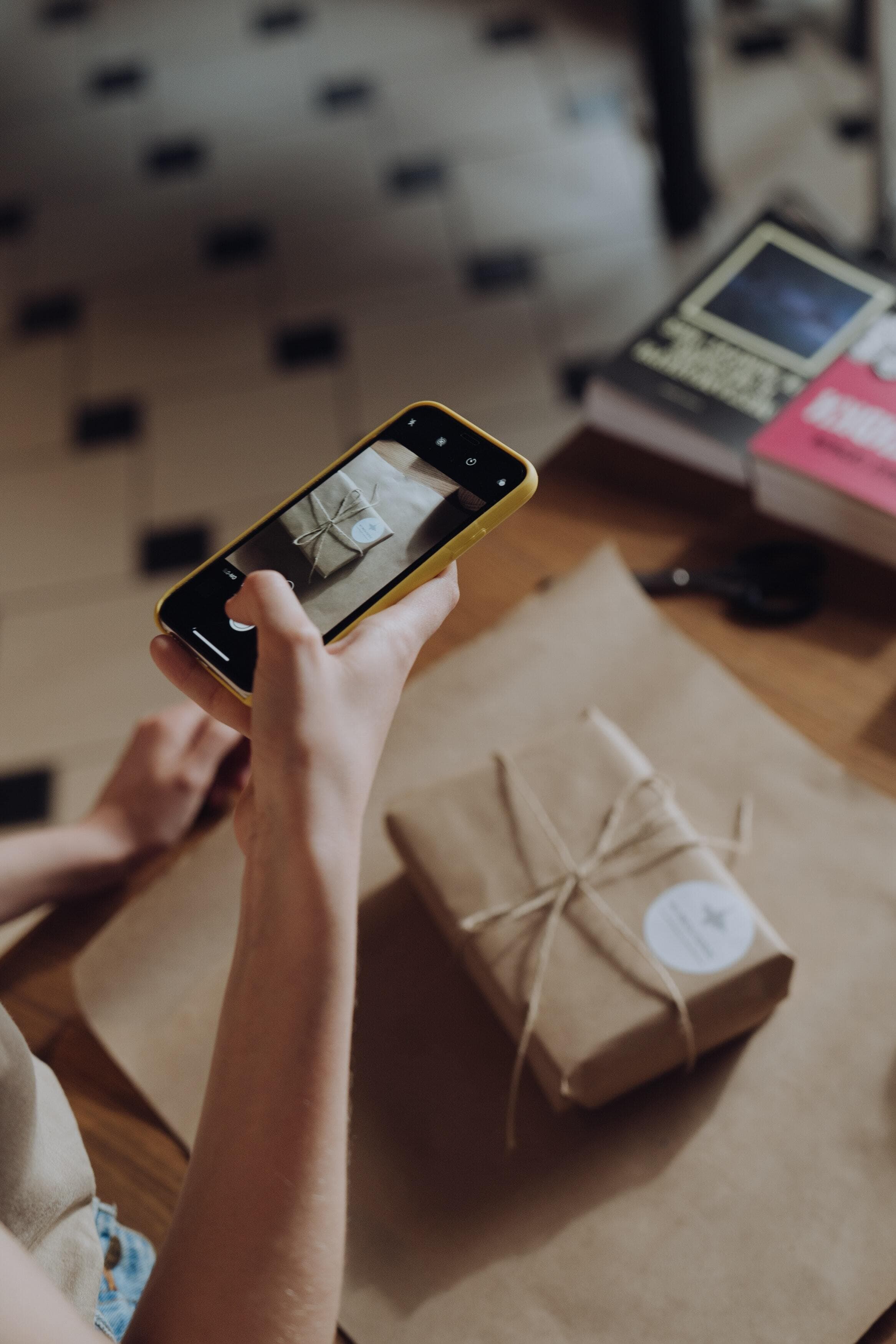 Someone is taking a picture of a gift wrapped in brown paper with a brown bow and a white, circular sticker on the top right corner of the package.