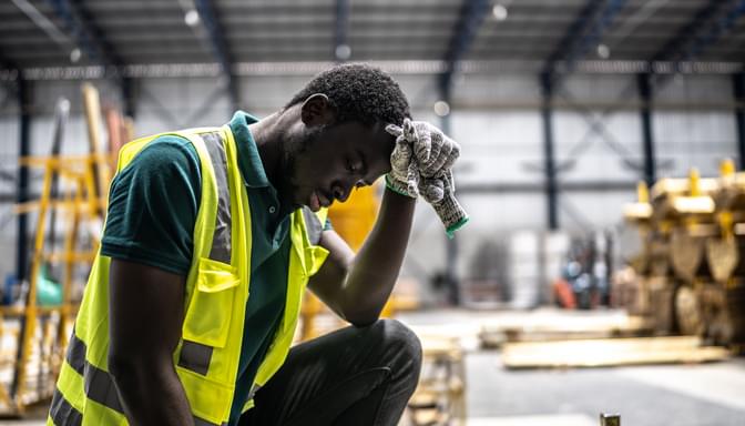A manufacturing employee is bent over his knee with a hand on his forehead out of mental exhaustion and is having issue with his mental health.