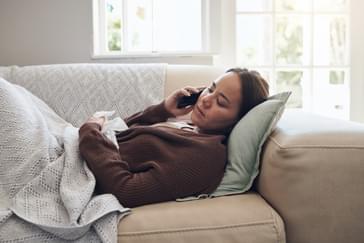 employee calling in sick to work laying on a couch holding a tissue in their left hand.