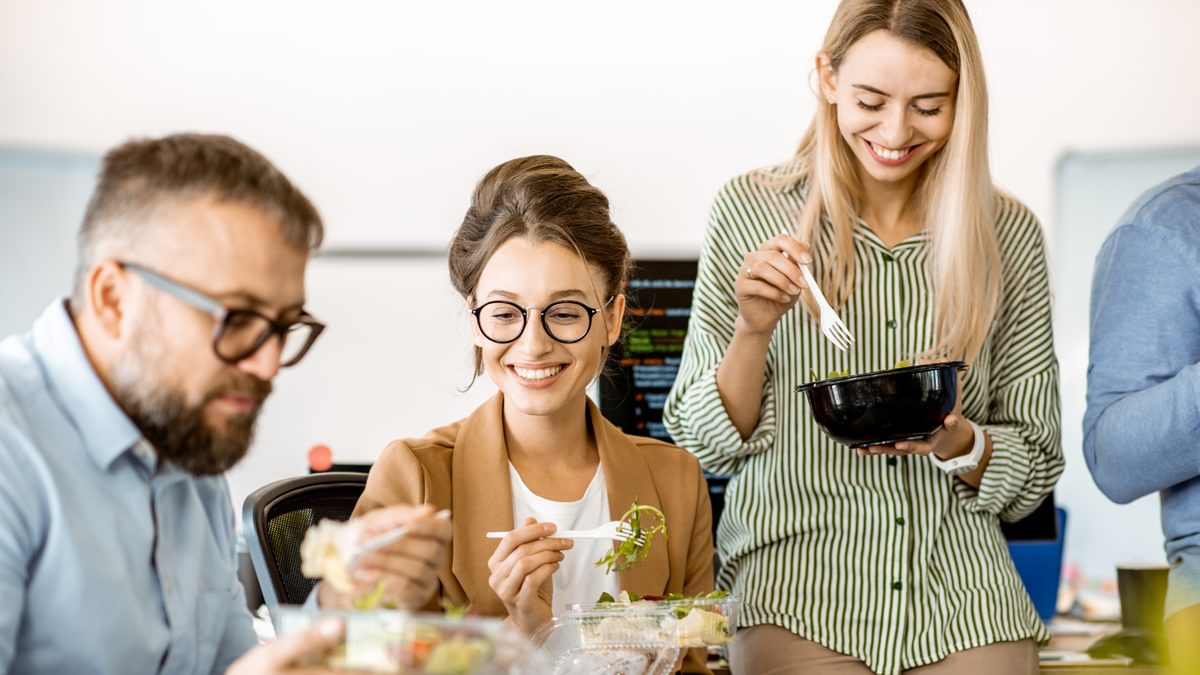 Drei Kollegen essen gemeinsam gesunde Mahlzeiten im Büro und lachen dabei.