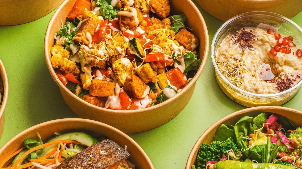 Eine Auswahl farbenfroher, frischer Catering-Bowls mit Gerichten wie gegrilltem Lachs mit Gemüse, Avocado-Salat, Tempeh- und Ofengemüse-Salat, Hummus mit Granatapfelkernen und verschiedenen bunten Toppings, arrangiert auf grünem Hintergrund.