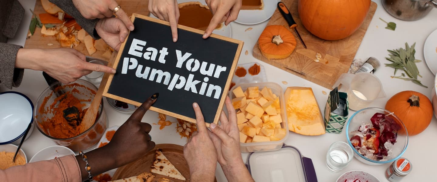 A tabletop with lots of different pumpkin food-items like pumpkin chunks, pumpkin dip, blitzed pumpkin in a pan... 7 hands are pointing to a sign in the middle saying 'Eat Your Pumpkin'.