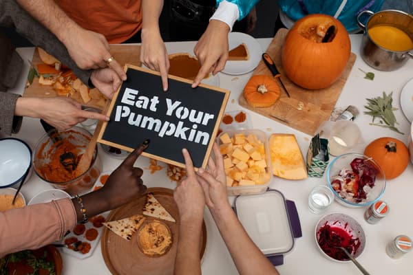 A tabletop with lots of different pumpkin food-items like pumpkin chunks, pumpkin dip, blitzed pumpkin in a pan... 7 hands are pointing to a sign in the middle saying 'Eat Your Pumpkin'.