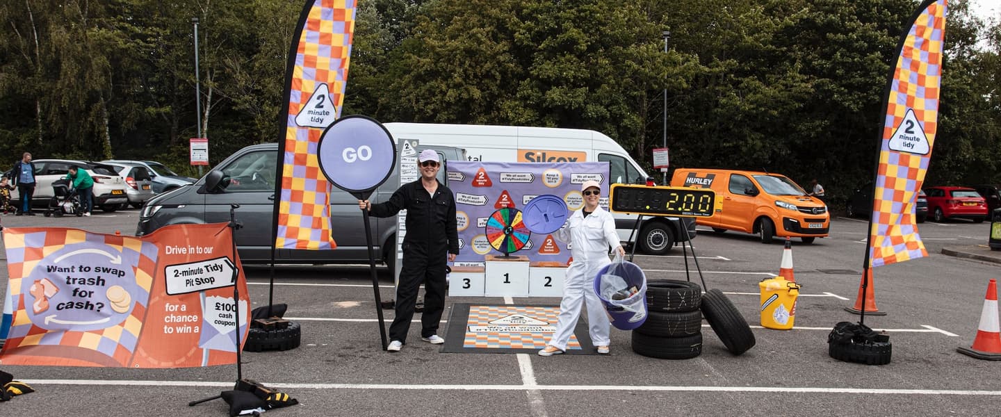 A colourful pit stop in the middle of a car park. There is a spin wheel and podium in the background. Two people wearing painters' jumpsuits stand facing the camera, one holding a 'go' sign.