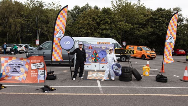 A colourful pit stop in the middle of a car park. There is a spin wheel and podium in the background. Two people wearing painters' jumpsuits stand facing the camera, one holding a 'go' sign.
