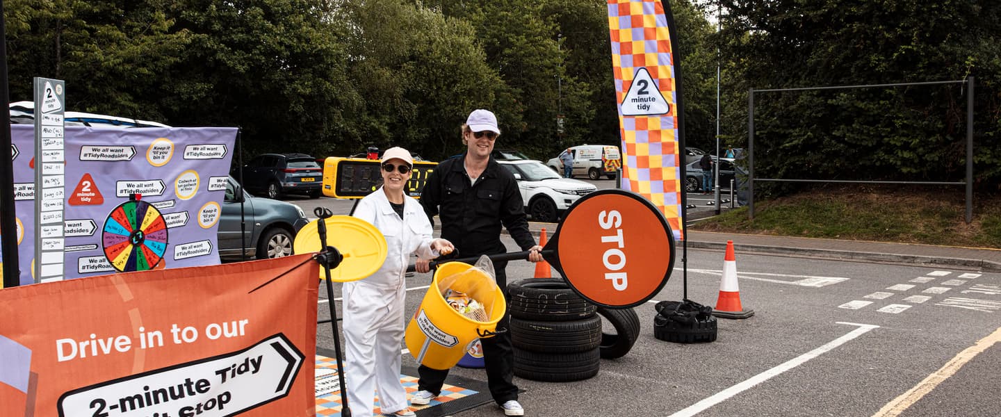 In a carpark, colourful signage signals a pit stop. A man and a woman hold a bin and a 'stop' sign in front of a space where a car can stop. Signage in bright orange and purple says '2 minute tidy' and 'drive in to our 2-minute Tidy Roadsides stop'
