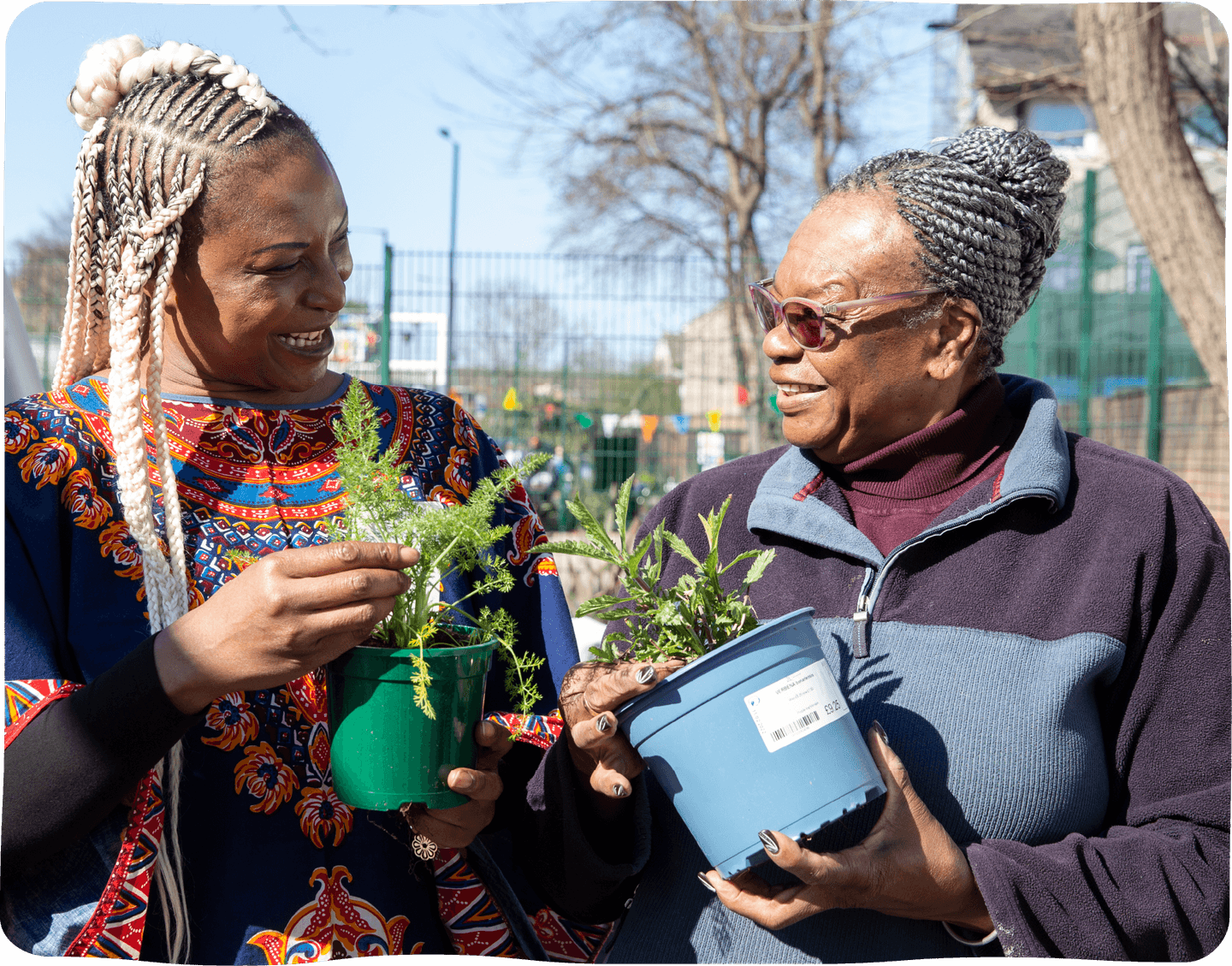 Two black women stand outside and lean in towards one an other, smiling as they hold potted plants.