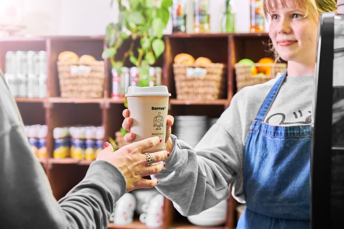 An apron-wearing young woman behind a till hands over a cup of coffee to a customer. The cup is a reusable cup, a beige colour with a dark green 'Borrow. Return for £1' written on the cup and an illustrated earth.