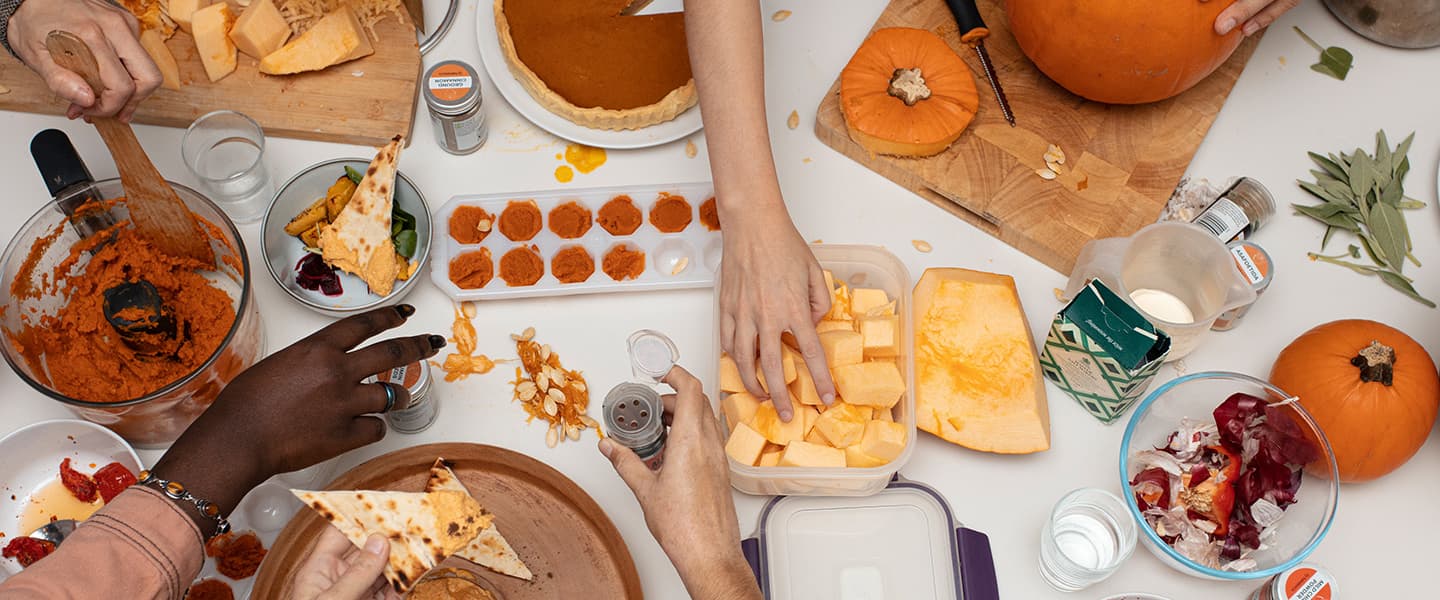 A table full of pumpkin goodies, with hands reaching from off-screen to grab things from the table. There are pumpkin pies, chopped pumpkin bits, pureed pumpkin in a muffin tin, herbs, flatbread...