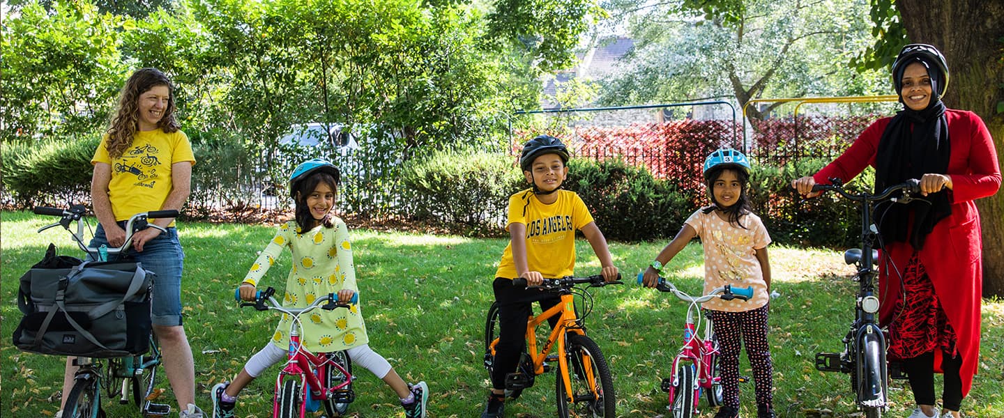 Two women and three children standing in a line with their bikes in a park on a sunny day.
