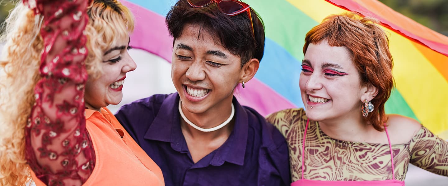 Three people smiling together in front of a Pride flag.