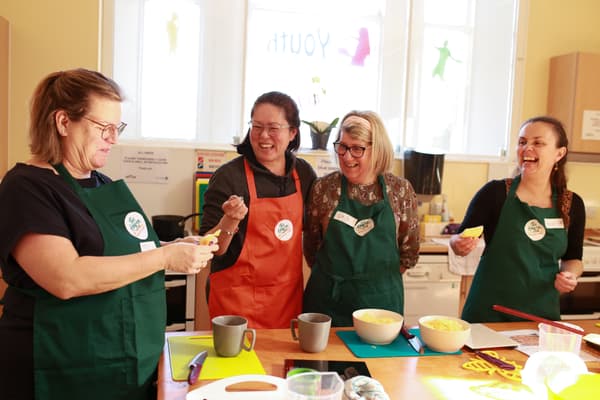 Four middle-aged women stand around a table, laughing. They are all wearing green aprons and one of the women is holding a lemon peel.
