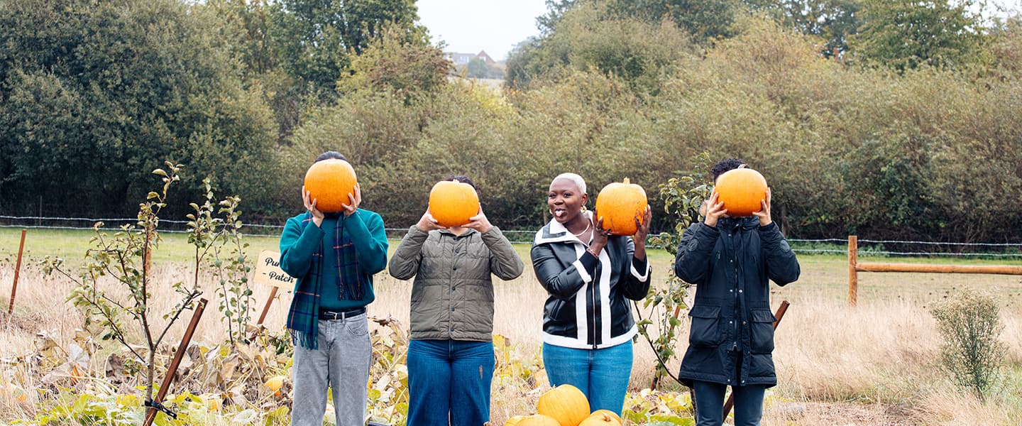 Four young adults stand in a field, behind a wheelbarrow filled with pumpkins. Three are holding a pumpkin in front of their faces, while one (a young black woman with short blonde hair) playfully sticks her tongue out and holds her pumpkin slightly away from her face.