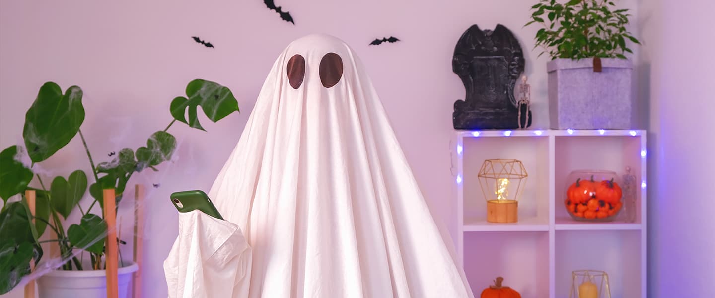 A person dressed up as a ghost in a white bedsheet stands facing the camera in a trendy bedroom, with plants and neon lights